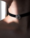 A close-up of a person's neck and shoulder wearing a black velvet choker necklace with a round silver-toned medallion centerpiece.