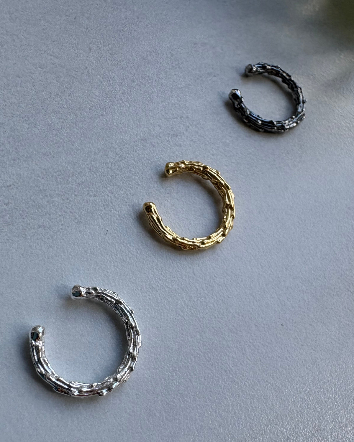 Three ear cuff earrings in black, gold, and silver on a gray surface.