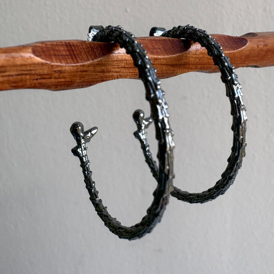 Two oxidised silver earrings on a wooden stick against a gray background
