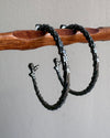 Two oxidised silver earrings on a wooden stick against a gray background