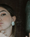 A woman with brown hair and winged eyeliner wearing a black tank top and a large silver textured earring, posing against a dark rustic wooden background.