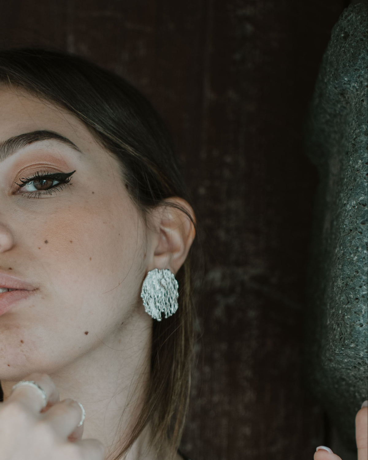 A woman with brown hair and winged eyeliner wearing a black tank top and a large silver textured earring, posing against a dark rustic wooden background.