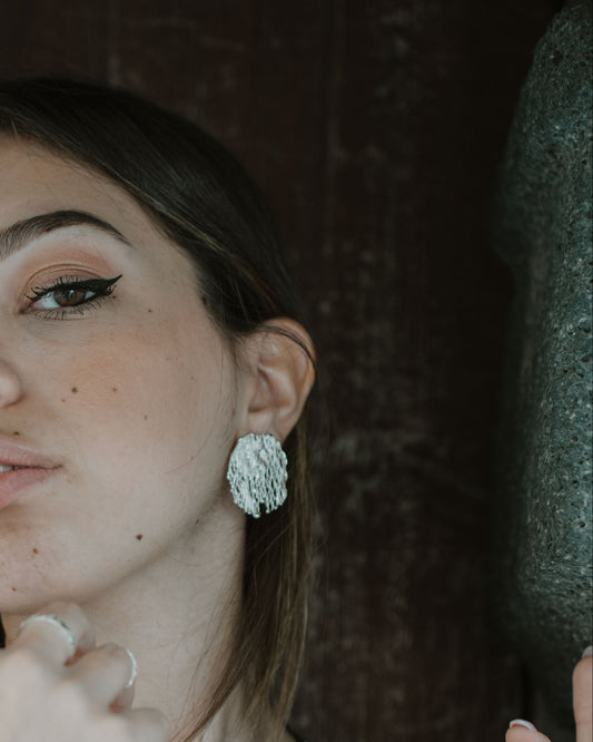 A woman with brown hair and winged eyeliner wearing a black tank top and a large silver textured earring, posing against a dark rustic wooden background.